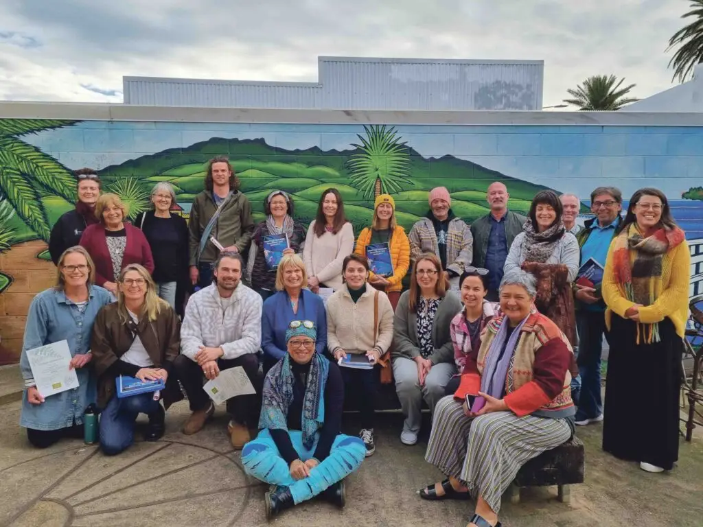 A diverse group of 25 participants poses in front of a colorful mural, holding documents, showcasing a collaborative learning environment.