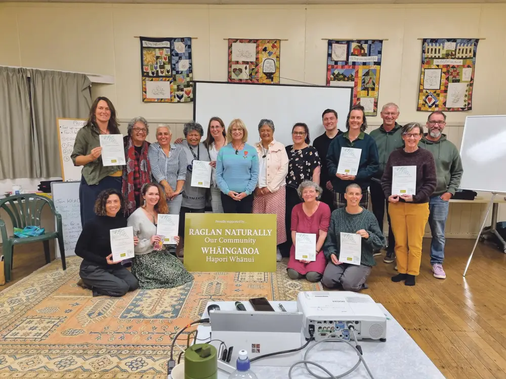 Group photo of diverse individuals holding certificates, gathered around a sign that reads "Raglan Naturally Our Community Whāingaroa." The setting features wooden floors and colorful wall hangings.