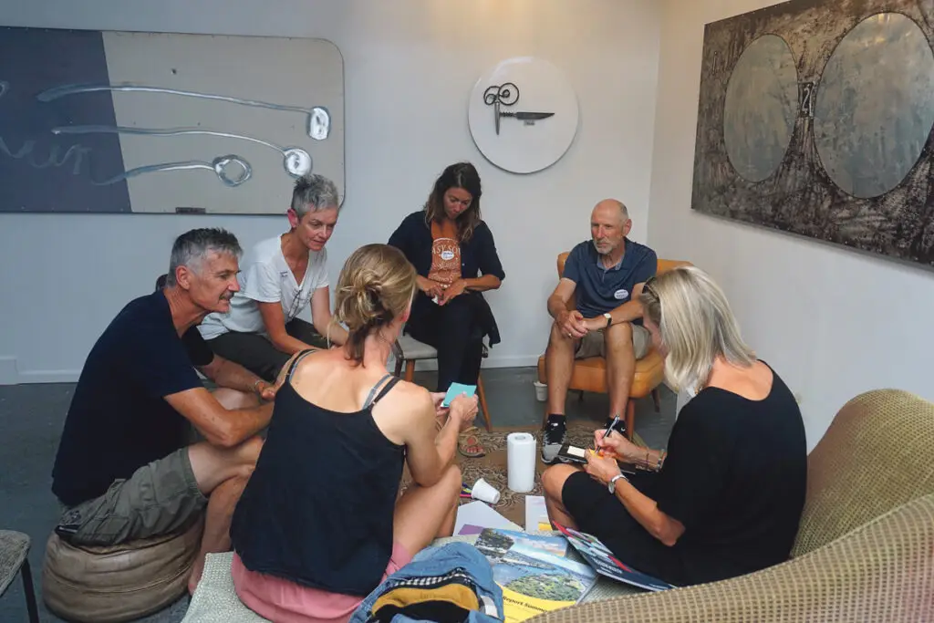 A group of six people engaged in a discussion around a coffee table, with art pieces on the walls and casual seating.