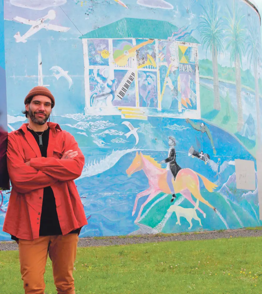 A man in a red shirt stands in front of a colorful mural featuring musical elements, horses, and waves, set in a vibrant landscape.