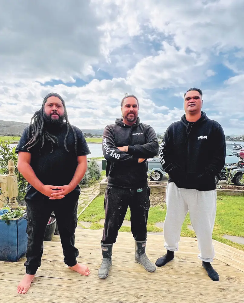 Three men stand on a wooden deck, wearing black hoodies and casual pants, against a backdrop of cloudy skies and green grass.