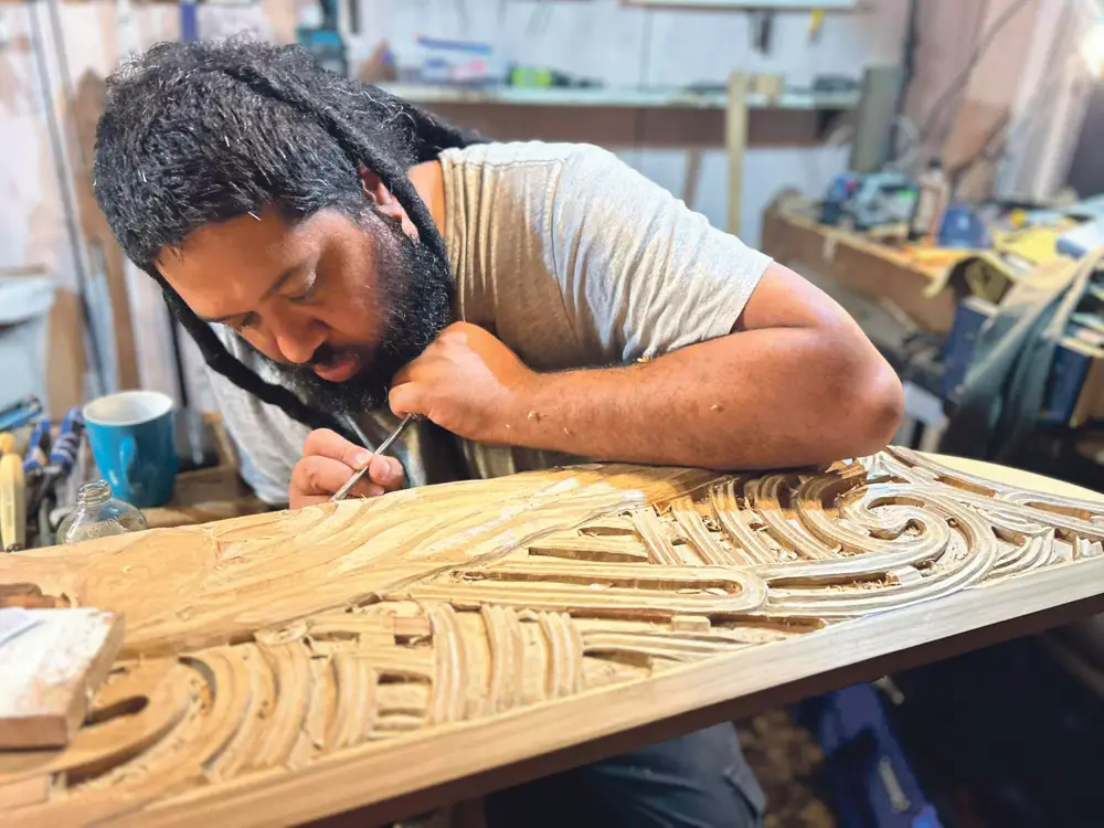 A craftsman with dreadlocks chisels intricate carvings on a wooden panel in a workshop, surrounded by tools and materials.