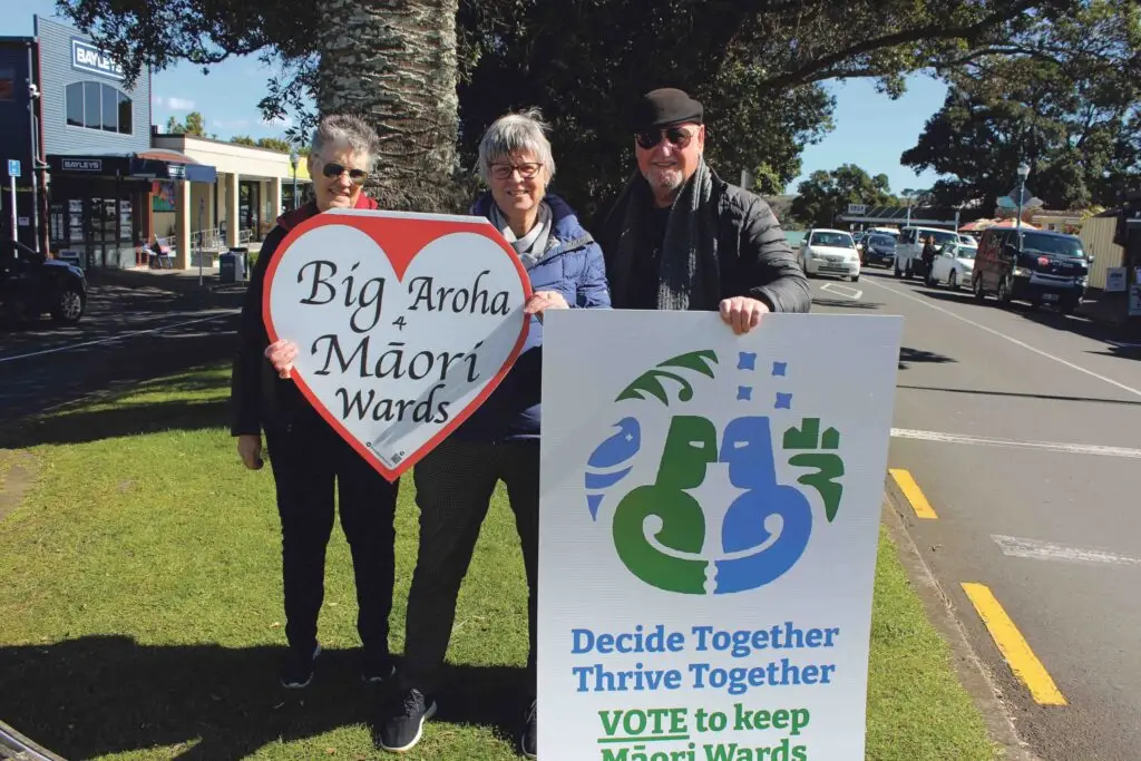 Three people stand on a street corner holding a large heart-shaped sign reading "Big Aroha for Māori Wards" and a poster advocating for Māori Wards.