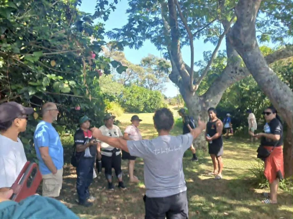 A group of people gather under trees in a lush green area, engaged in discussion and activities related to community initiatives.