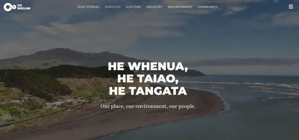 Coastal landscape of Raglan with rolling hills, ocean waves, and a beach, featuring the text "HE WHENUAS, HE TAIAO, HE TANGATA" prominently.