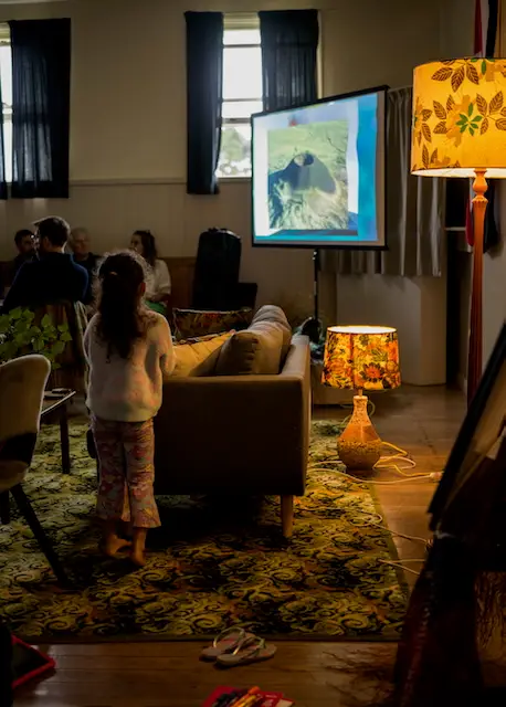 A child stands in front of a projector displaying Karioi Volcano, with a cozy room featuring a patterned rug, sofa, and decorative lamp.