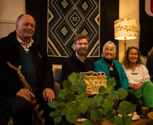 Group of four individuals seated together, with varied expressions, surrounded by a plant and patterned decor, highlighting a community gathering.