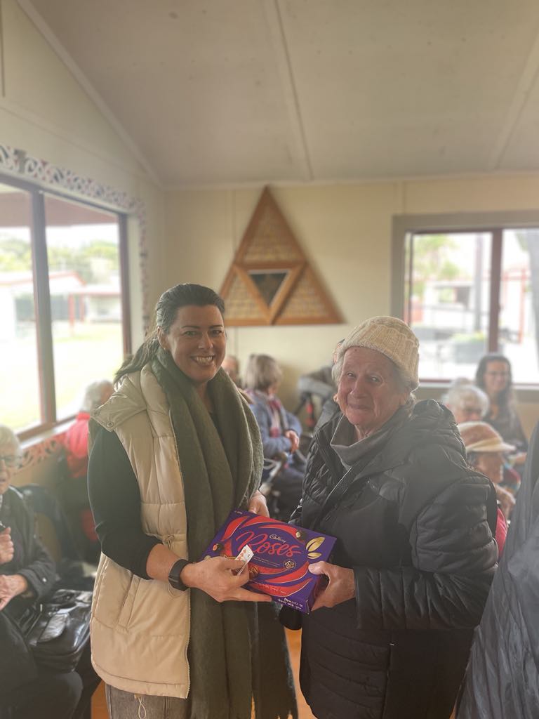 A smiling woman hands a box of chocolates to an elderly woman in a warm, inviting room with other people seated in the background.