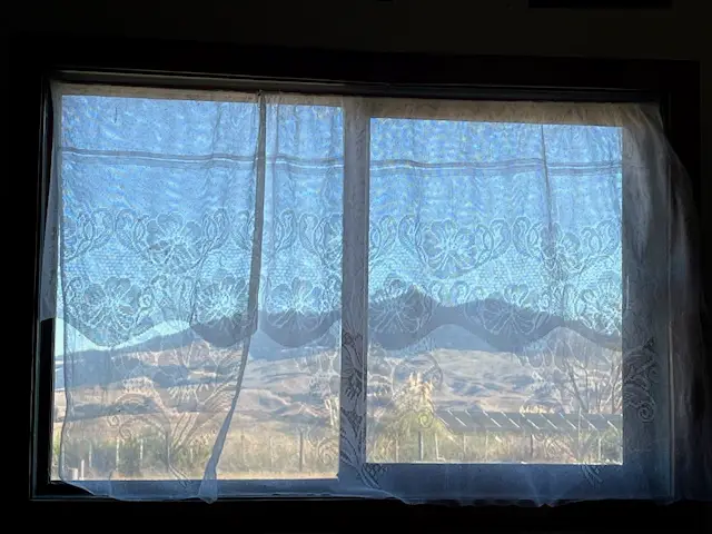 Sheer lace curtains frame a window revealing a picturesque landscape of rolling hills under a clear blue sky.