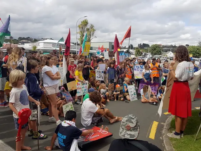 A diverse group of children and adults gathered on a street, holding colorful signs and flags, engaging in a community protest or rally.