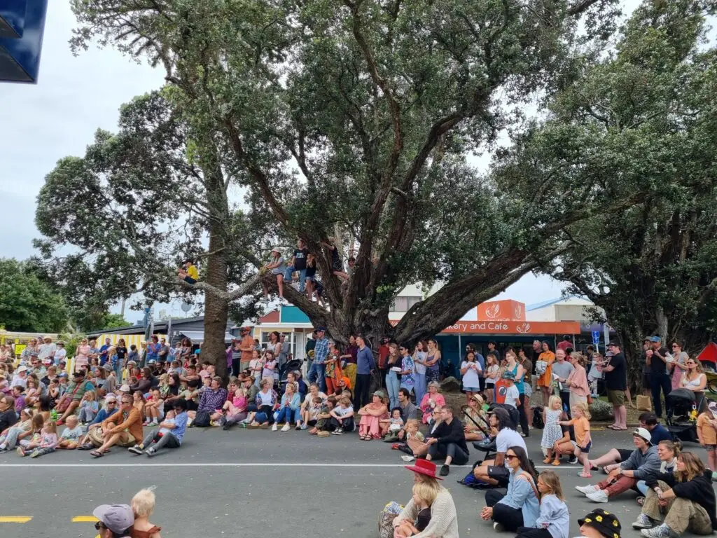 A crowd of diverse people gathers under a large tree, watching an event in a town setting, with colorful shop fronts in the background.