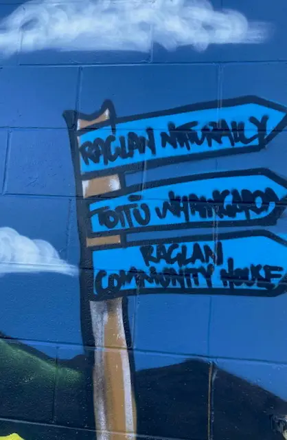 Colorful mural featuring wooden signposts with "Raglan Naturally," "Toitū Whāingaroa," and "Raglan Community House" written in bold letters.