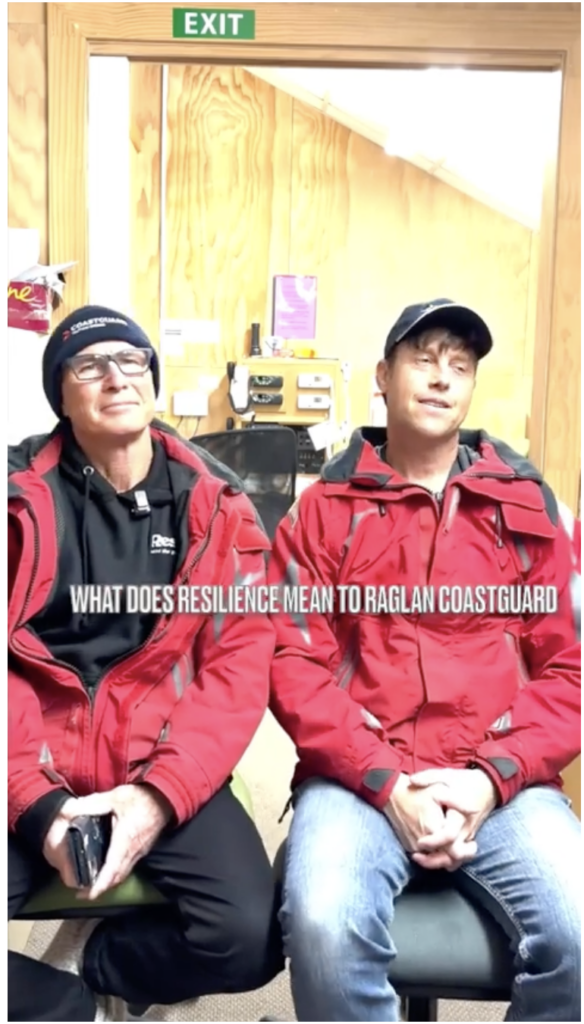 Two individuals in red jackets sit in an indoor space, looking relaxed, with a sign above them reading "EXIT." Text asks, "What does resilience mean to Raglan Coastguard?"