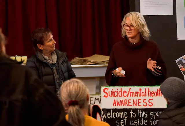 Two women engage in discussion at a community event, with a sign promoting suicide and mental health awareness in the foreground.
