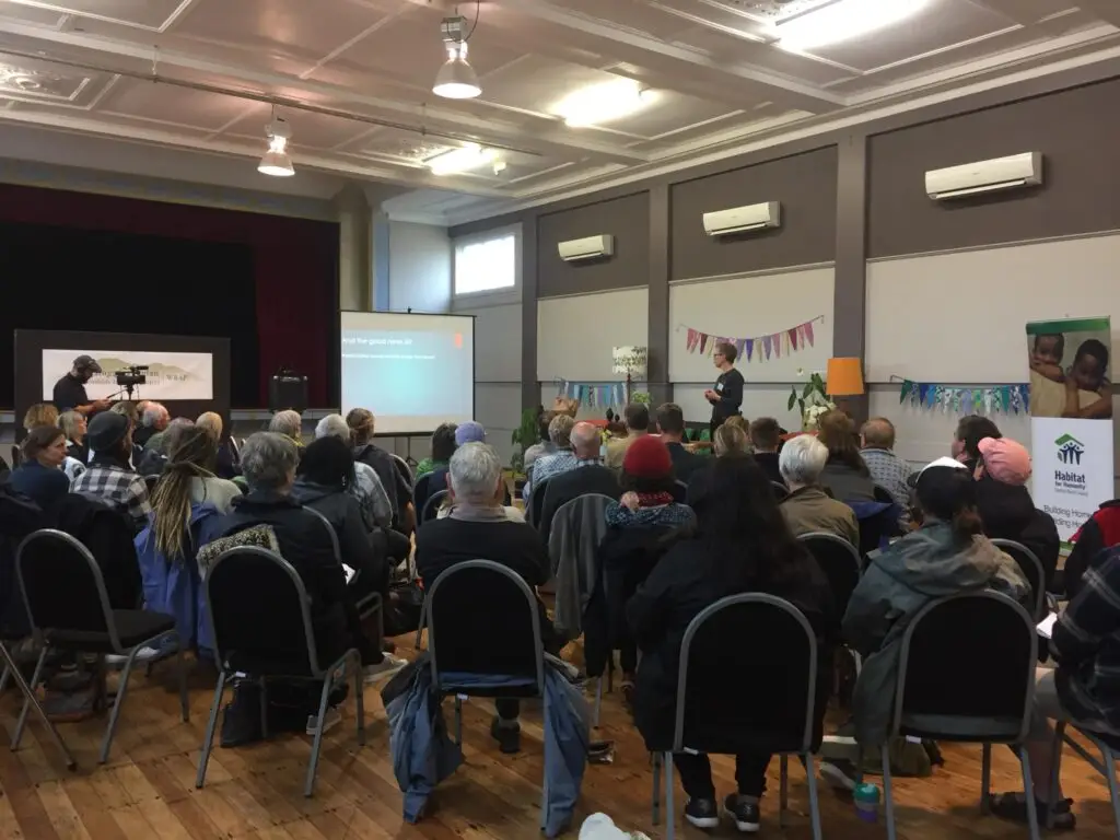 Community meeting in a hall with a speaker presenting on a screen, surrounded by an audience seated in rows, colorful bunting in the background.