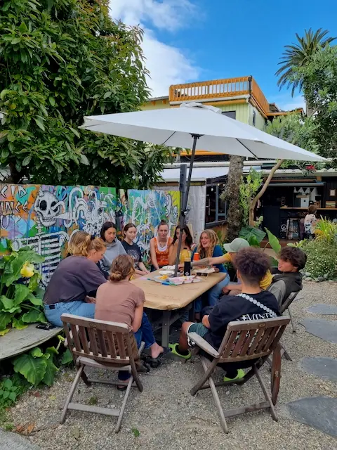 A group of young people gathers around a large table under an umbrella, surrounded by greenery and colorful murals in a casual outdoor setting.